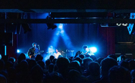 A lively concert scene with a band performing on stage under blue lights. The audience is packed, creating an energetic atmosphere in the dark venue.