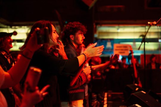 Audience members applaud enthusiastically at a dimly lit concert, with warm red lighting. A microphone stand is visible, highlighting a lively atmosphere.