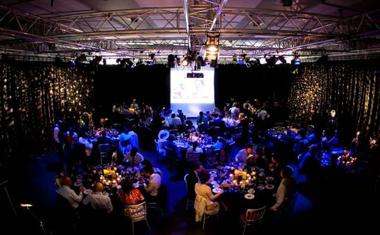 A dimly lit banquet hall with round tables, elegantly decorated with candles. Guests sit and converse. A projector displays images on a large screen.