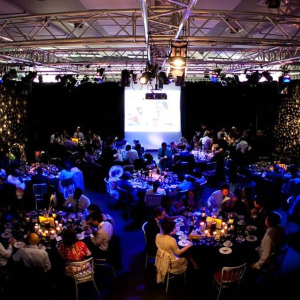 A dimly lit banquet hall with round tables, elegantly decorated with candles. Guests sit and converse. A projector displays images on a large screen.
