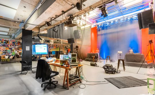 Spacious studio with equipment, computers, and cameras. Red and blue lighting create a vibrant atmosphere. Walls adorned with colorful posters.