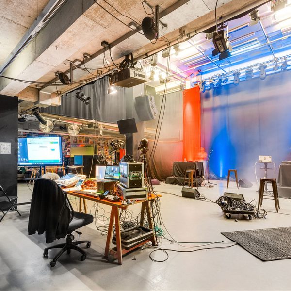 Spacious studio with equipment, computers, and cameras. Red and blue lighting create a vibrant atmosphere. Walls adorned with colorful posters.