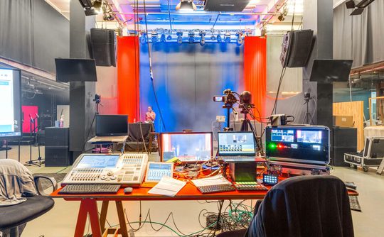 A vibrant TV studio with a red and blue lit stage. A control desk with monitors, soundboards, and cables dominates the foreground, suggesting a dynamic production environment.