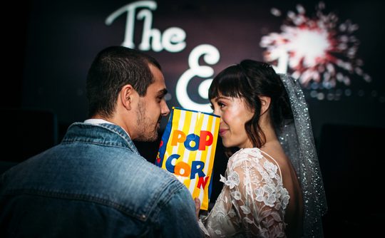 A bride and groom share popcorn, smiling intimately in a theater. Background shows "The End" and a firework, suggesting a joyful, cinematic theme.