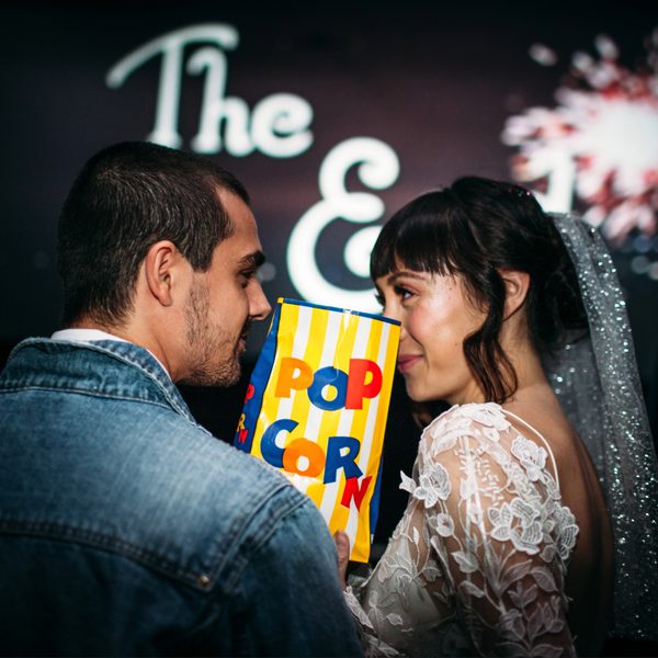 A bride and groom share popcorn, smiling intimately in a theater. Background shows "The End" and a firework, suggesting a joyful, cinematic theme.