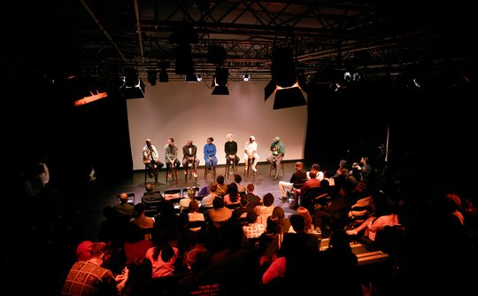 A panel of seven people sits on a dimly lit stage, facing an attentive audience in a theatre. The atmosphere is formal and focused, suggesting a discussion or Q&A session.