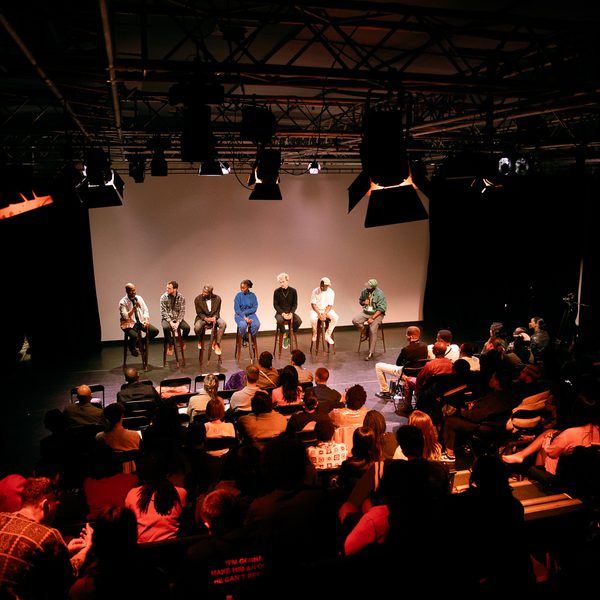 A panel of seven people sits on a dimly lit stage, facing an attentive audience in a theatre. The atmosphere is formal and focused, suggesting a discussion or Q&A session.
