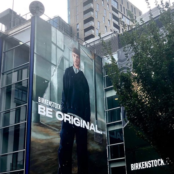 Billboard on a city building reads "Birkenstock: Be Original," showing a person in casual attire. The backdrop features urban high-rises and trees.