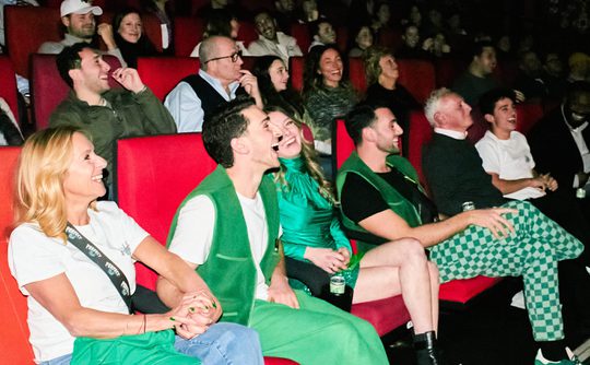 A group of people sit in red theatre seats, laughing and enjoying a show. The mood is lively and cheerful, with a focus on joy and camaraderie.