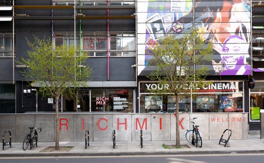 Exterior of Rich Mix cultural center, featuring colourful mural, trees, and bicycles parked by the entrance. The atmosphere is vibrant and inviting.