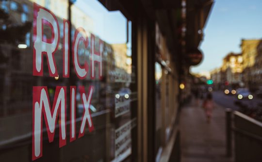 Reflective window with "Rich Mix" in bold red letters, blurred urban street scene, warm evening light creating a vibrant, bustling atmosphere.