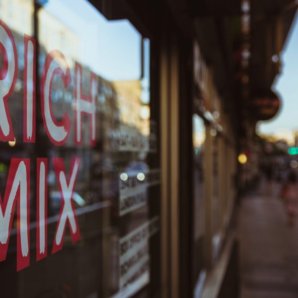 Reflective window with "Rich Mix" in bold red letters, blurred urban street scene, warm evening light creating a vibrant, bustling atmosphere.