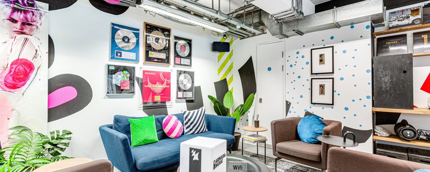 Vibrant lounge with colorful decor, including blue sofas with pink and green cushions. Walls display music records and abstract art, creating a lively atmosphere.