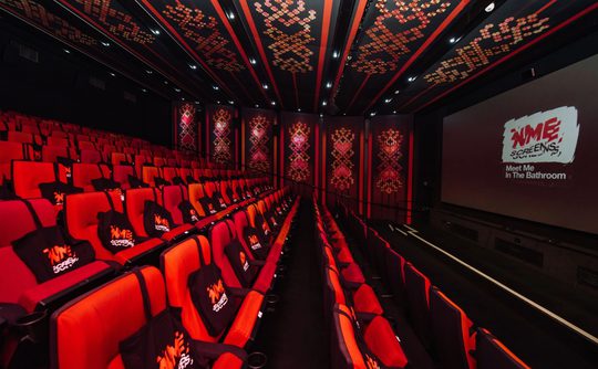 A stylish cinema screen with red and black decor, featuring rows of empty red seats with black tote bags. A large screen displays "NME Screens: Meet Me in the Bathroom."
