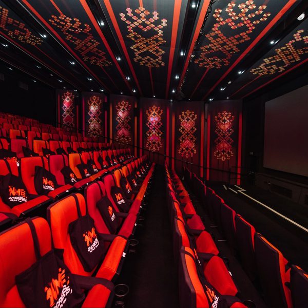 A stylish cinema screen with red and black decor, featuring rows of empty red seats with black tote bags. A large screen displays "NME Screens: Meet Me in the Bathroom."