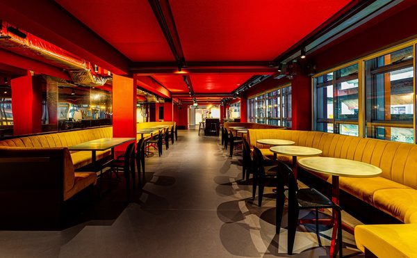 A modern restaurant interior features long yellow booths, black tables and chairs, and bright red ceiling panels. The empty dining room feels warm and orderly.