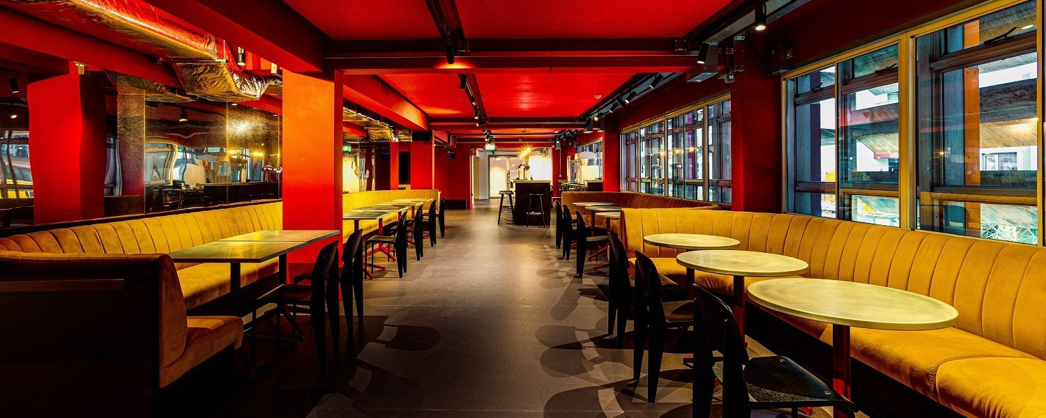 A cafe interior with vibrant red walls and ceiling, yellow booth seating, and wooden tables. Large windows line one side, creating a warm, inviting atmosphere.
