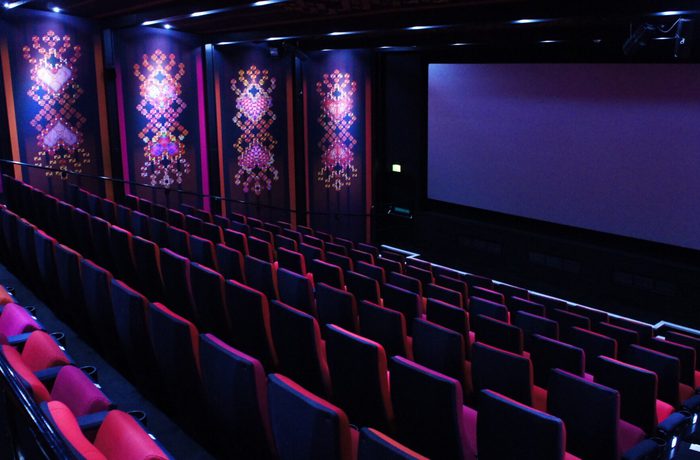 An empty movie theater with rows of red and black seats facing a large blank screen. The walls feature colourful geometric patterns under dim lighting, creating an intimate atmosphere.