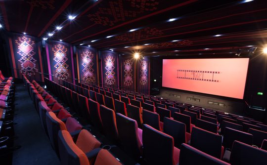 A brightly lit, empty cinema with red and purple patterned walls, plush red and purple seats, and a large screen displaying a filmstrip design.
