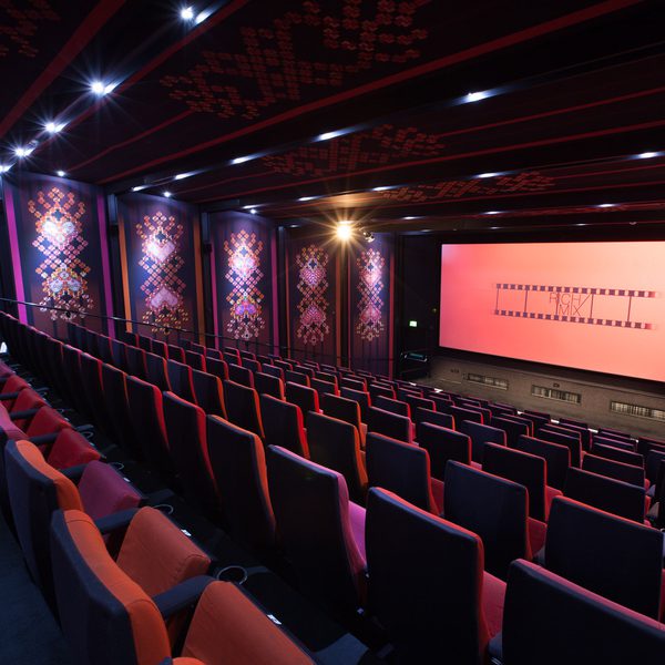 A brightly lit, empty cinema with red and purple patterned walls, plush red and purple seats, and a large screen displaying a filmstrip design.