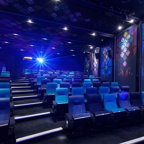 Empty movie theater with rows of alternating blue and dark blue seats, vibrant geometric wall art, and a bright blue projector light beaming.
