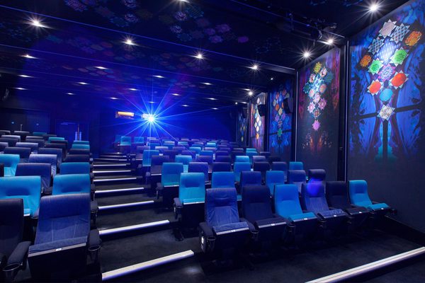Empty movie theater with rows of alternating blue and dark blue seats, vibrant geometric wall art, and a bright blue projector light beaming.