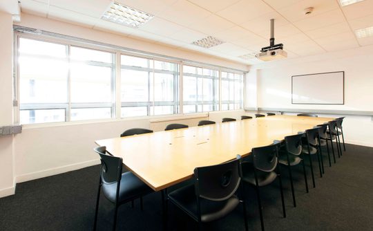 A well-lit meeting room with a long wooden table surrounded by black chairs. Large windows on the left allow natural light, creating a professional atmosphere.
