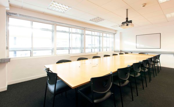 A well-lit meeting room with a long wooden table surrounded by black chairs. Large windows on the left allow natural light, creating a professional atmosphere.