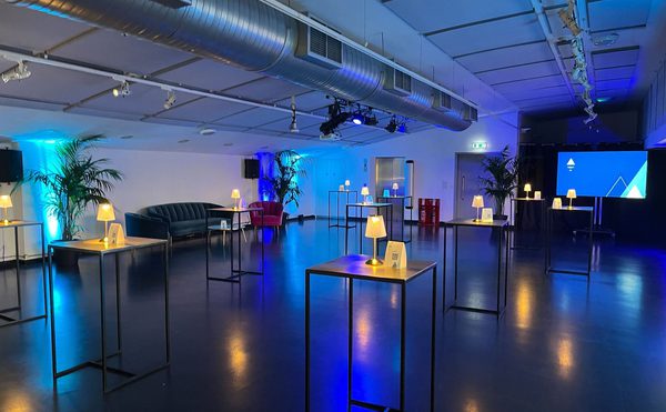 A modern conference room with high ceilings features tall tables and small lamps. Ambient blue lighting highlights the sleek, minimalist environment.