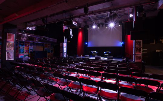 Dimly lit theatre with rows of empty red chairs facing a stage. The stage features a large screen and two black chairs, evoking an expectant atmosphere.