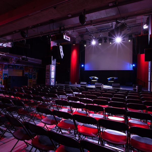 Dimly lit theatre with rows of empty red chairs facing a stage. The stage features a large screen and two black chairs, evoking an expectant atmosphere.
