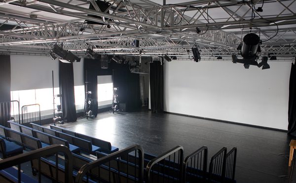 A small, empty theater features tiered blue seating facing a clear stage. Overhead, a grid of lighting fixtures hangs, with a large white wall at the back.