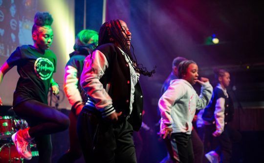 A group of dancers energetically performing on a stage under vibrant purple and green lights, wearing casual sporty outfits and jackets.