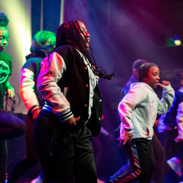 A group of dancers energetically performing on a stage under vibrant purple and green lights, wearing casual sporty outfits and jackets.