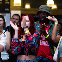 A diverse group of people enthusiastically clapping at a lively event. They wear casual, colorful attire, looking engaged and happy under warm lighting.