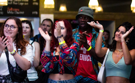 A diverse group of people enthusiastically clapping at a lively event. They wear casual, colorful attire, looking engaged and happy under warm lighting.