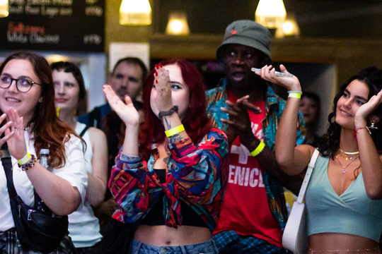 A diverse group of people enthusiastically clapping at a lively event. They wear casual, colorful attire, looking engaged and happy under warm lighting.