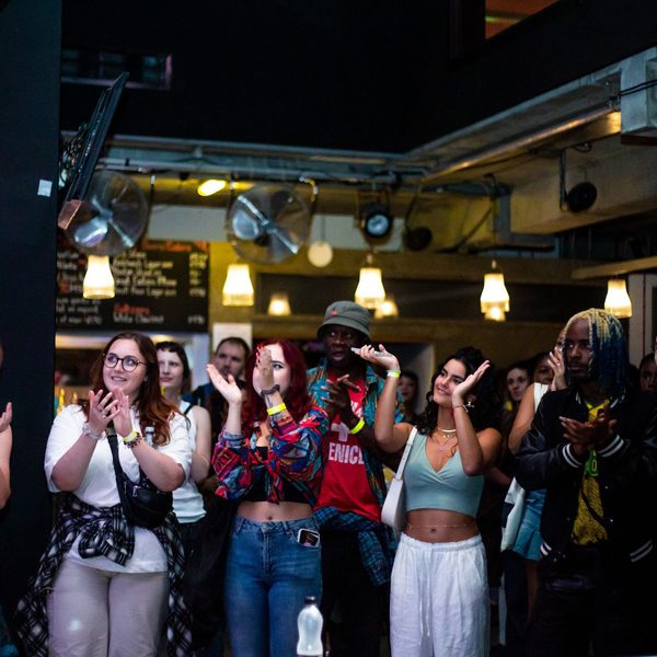 A diverse group of people claps and smiles in a lively indoor venue. Warm lighting and casual attire create an atmosphere of excitement and enjoyment.