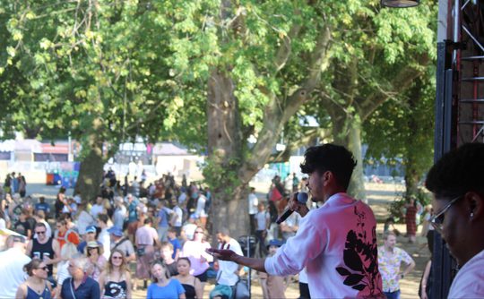 A performer in a white shirt with floral print sings into a microphone on an outdoor stage. A large crowd watches among trees, creating a lively festival atmosphere.