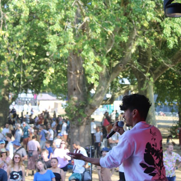 A performer in a white shirt with floral print sings into a microphone on an outdoor stage. A large crowd watches among trees, creating a lively festival atmosphere.