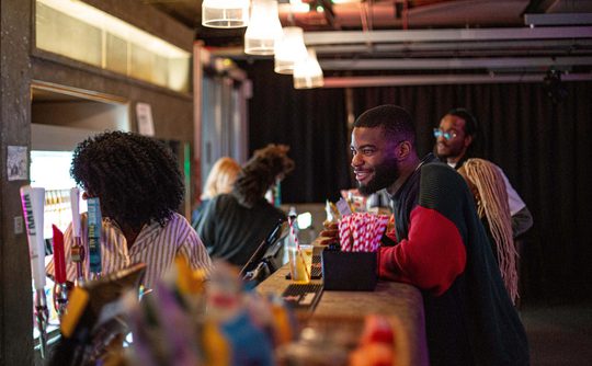 A person in a red and black sweater smiles while leaning on a bar counter with drinks and popcorn. Others chat nearby. The scene is warmly lit and lively.