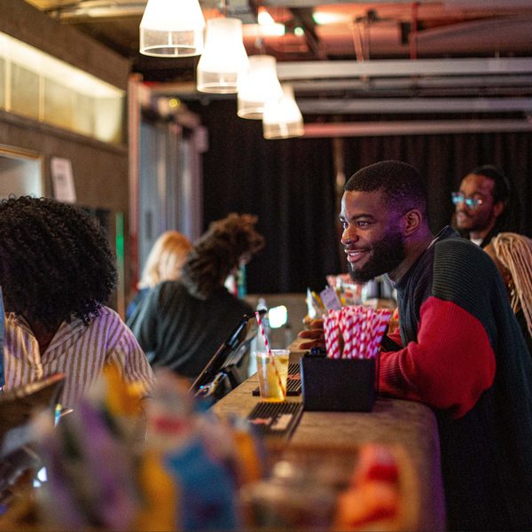 A person in a red and black sweater smiles while leaning on a bar counter with drinks and popcorn. Others chat nearby. The scene is warmly lit and lively.