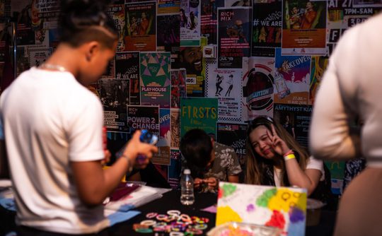A lively event scene with a person in focus creating crafts at a colourful, poster-covered table. Two people in the background are smiling energetically.