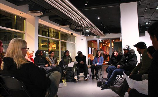 A diverse group of people sit in a circle in a modern, well-lit room, attentively sharing ideas. The setting is casual yet focused and collaborative.